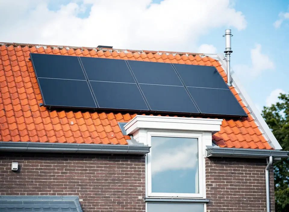 Acht zwarte zonnepanelen op het oranje pannendak van een bakstenen rijtjeshuis, boven een witte dakkapel